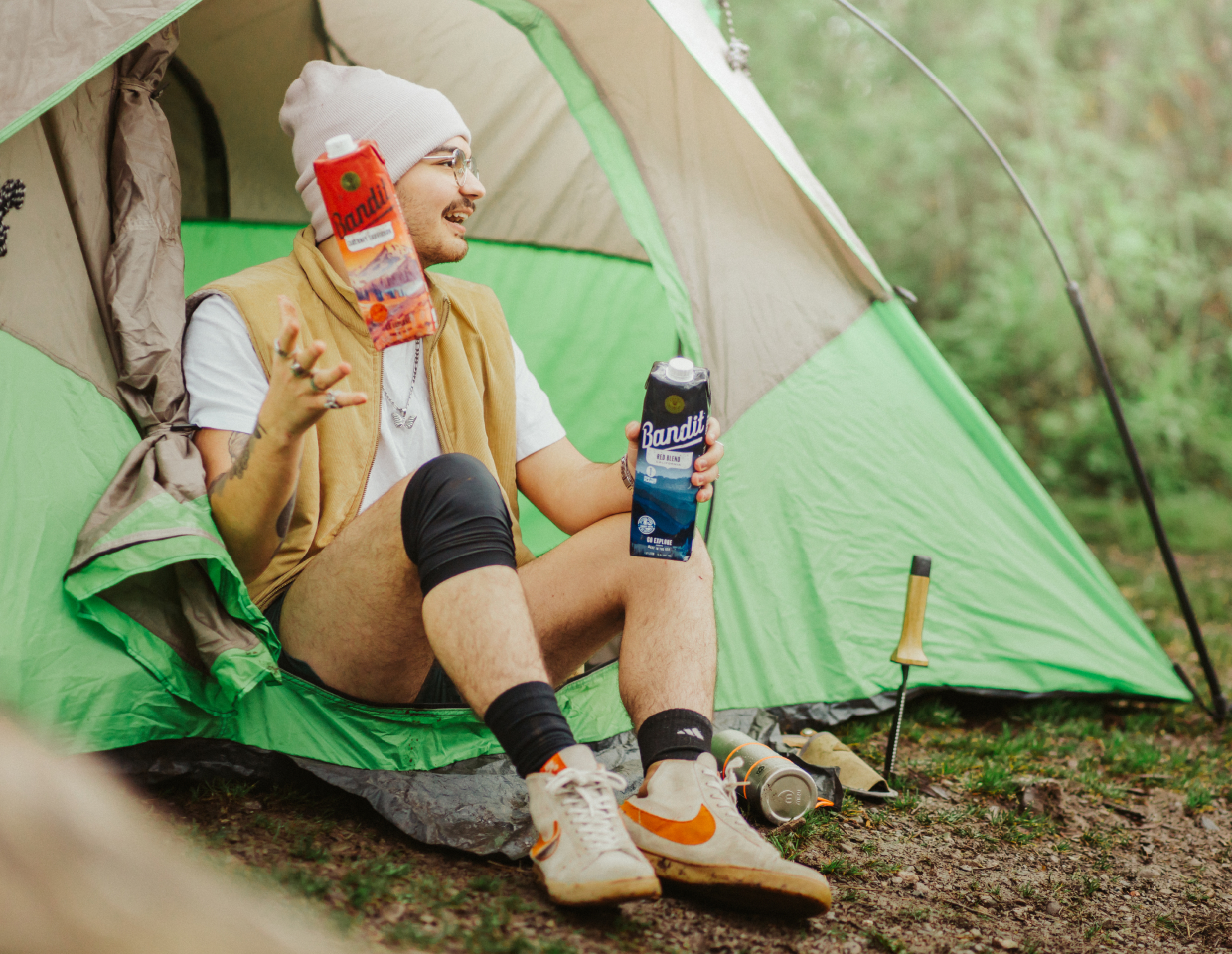 Hip young man camping in the forest and sitting in a tent holding two Bandit wines.