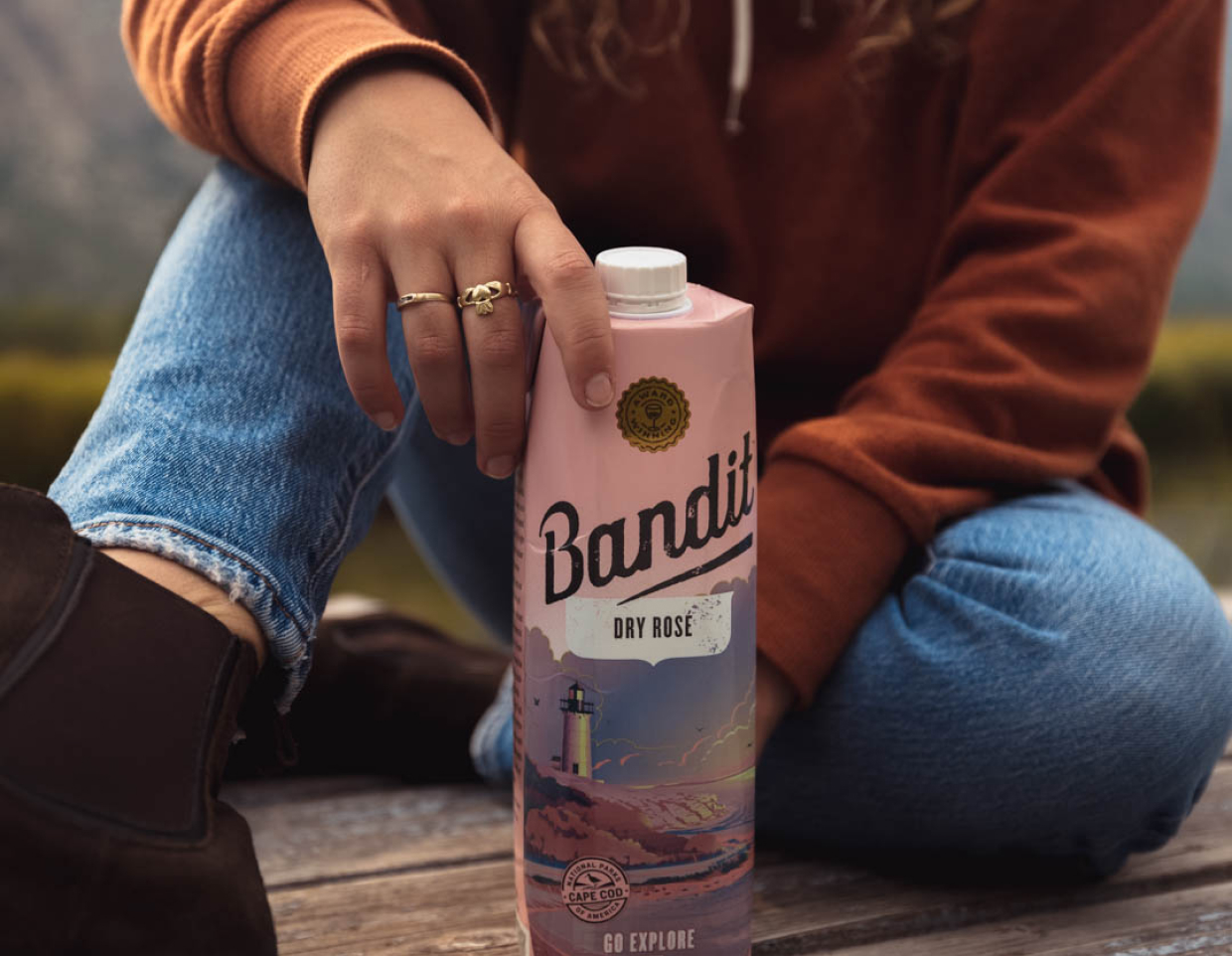 Closeup of a woman in a brown hoodie sitting on a dock holding a tetra pak of Bandit rose wine.