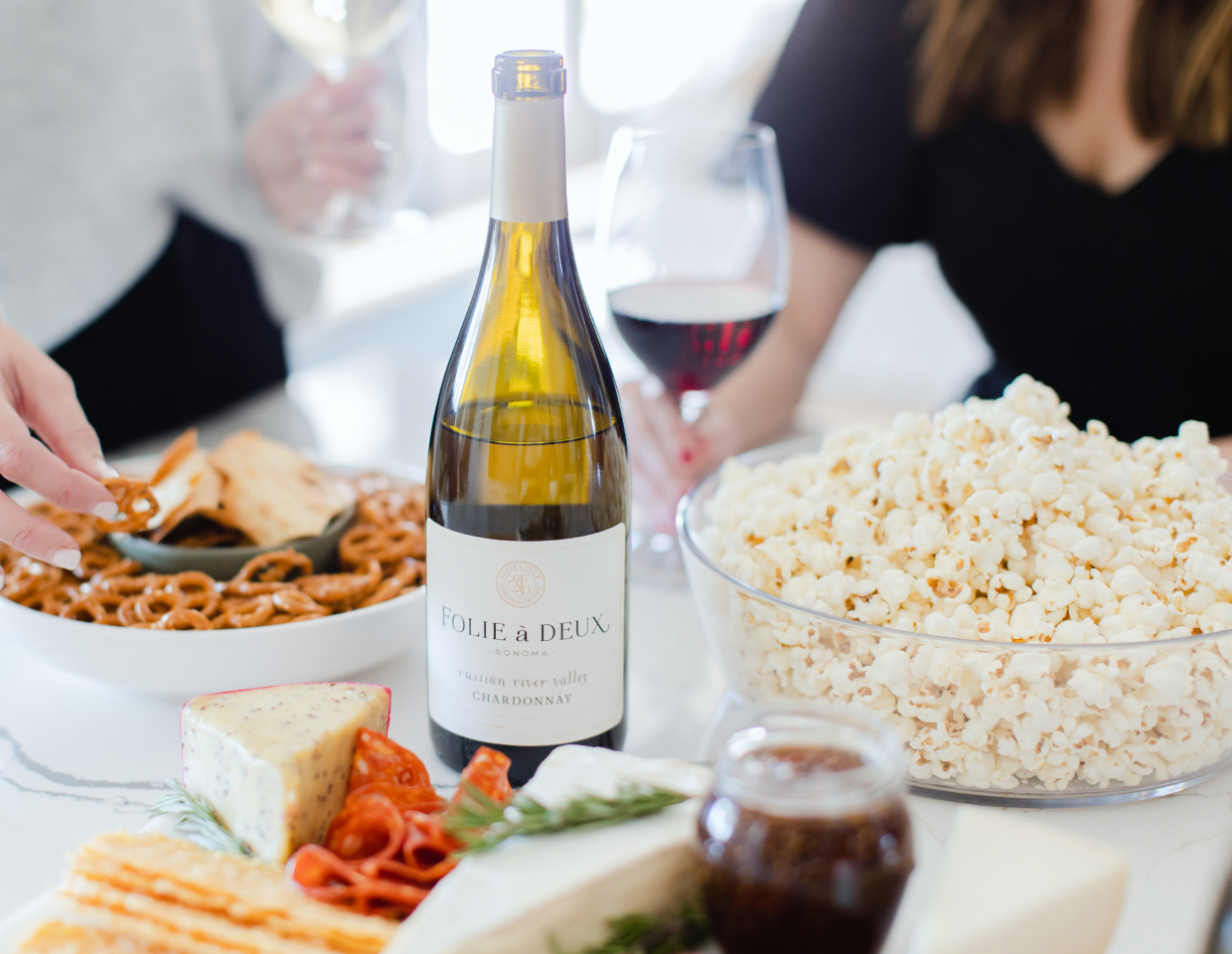 Bottle of Folie a Deux Chardonnay on a table full of snacks with people in the background.