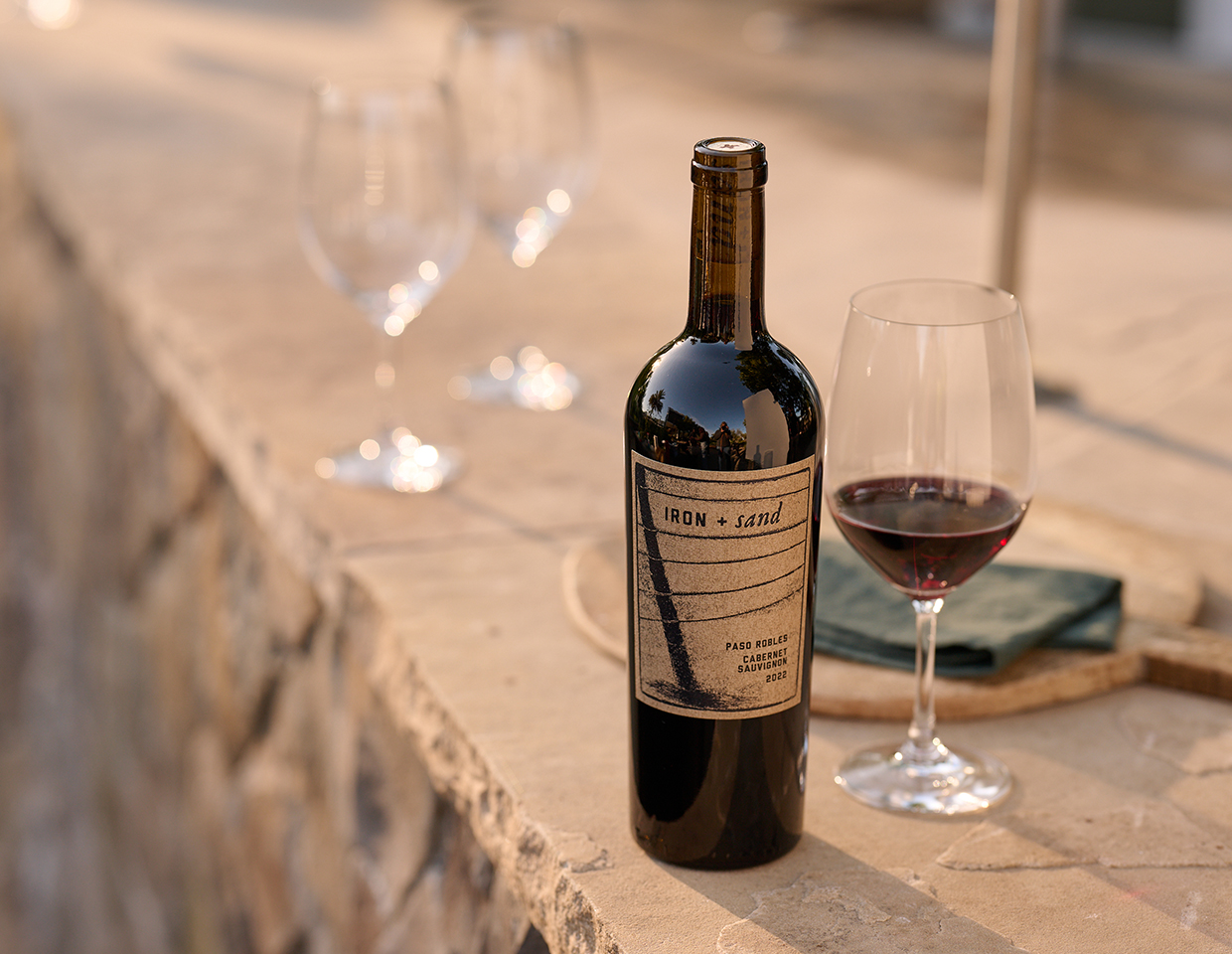 Bottle of Iron + Sand paso robles cabernet sauvignon vintage 2022 next to a glass of wine on a brown rock surface.