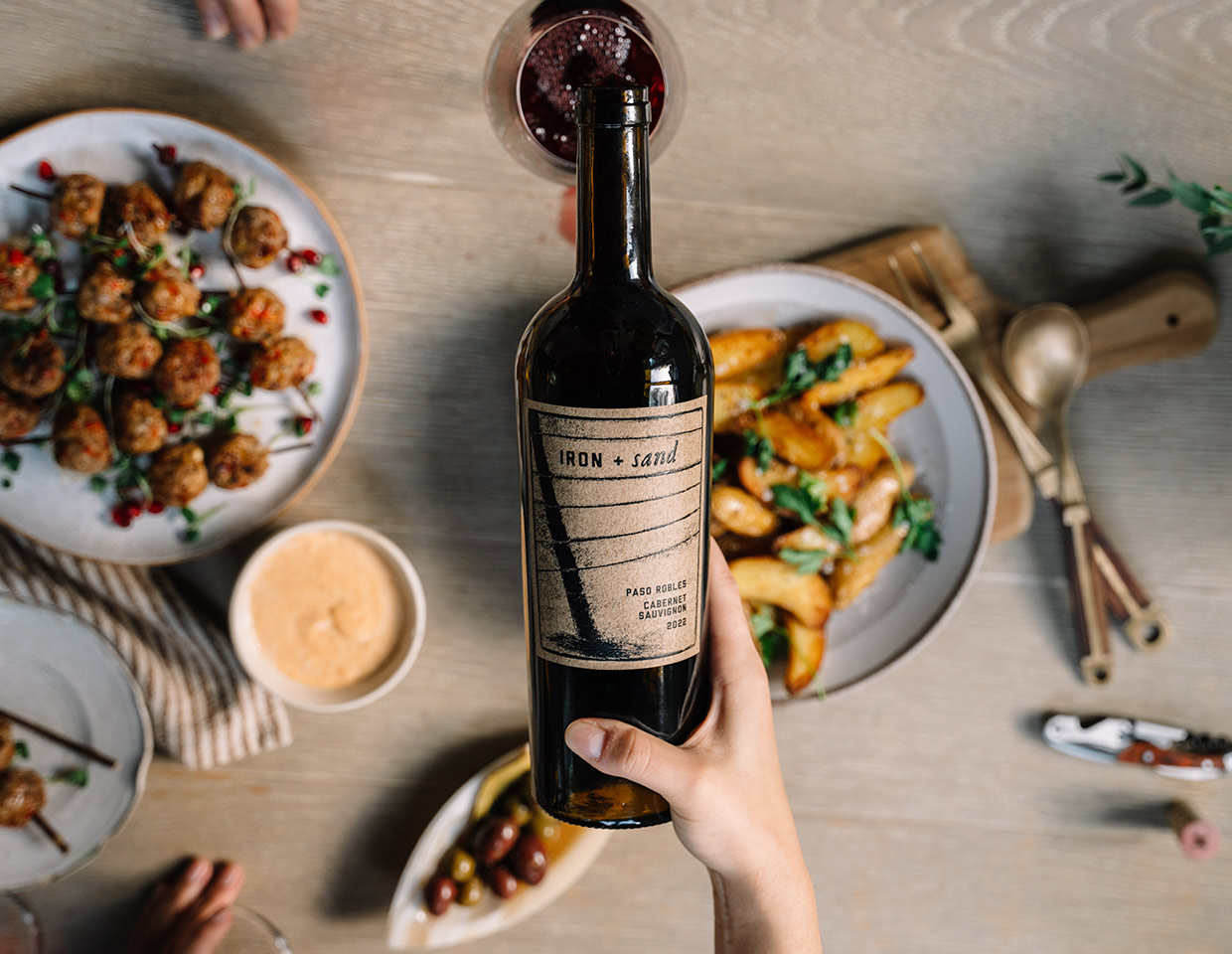 Person pouring a bottle of iron + Sand into a glass on a table set with appetizers