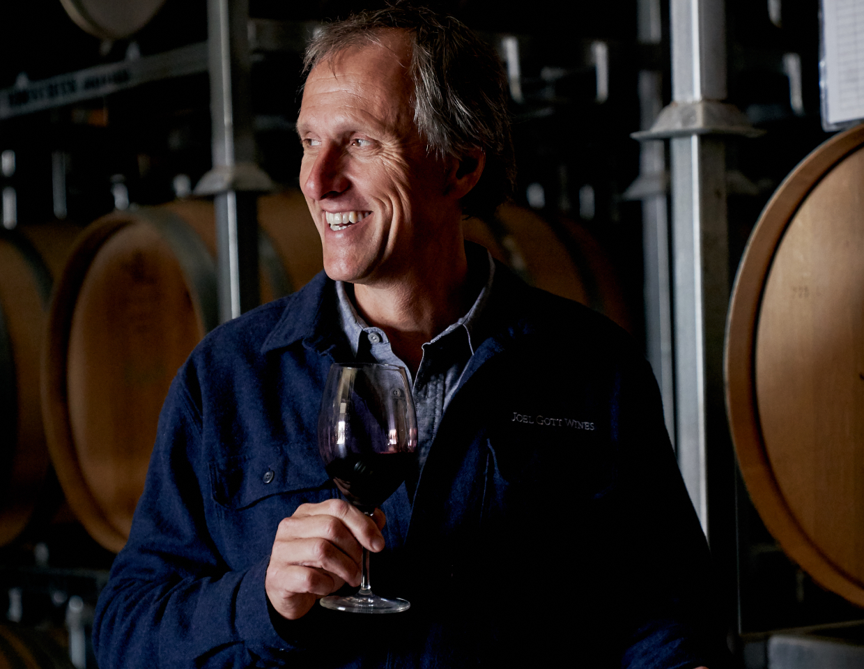 Joel Gott smiling and sampling wine in front of a row of wine barrels.