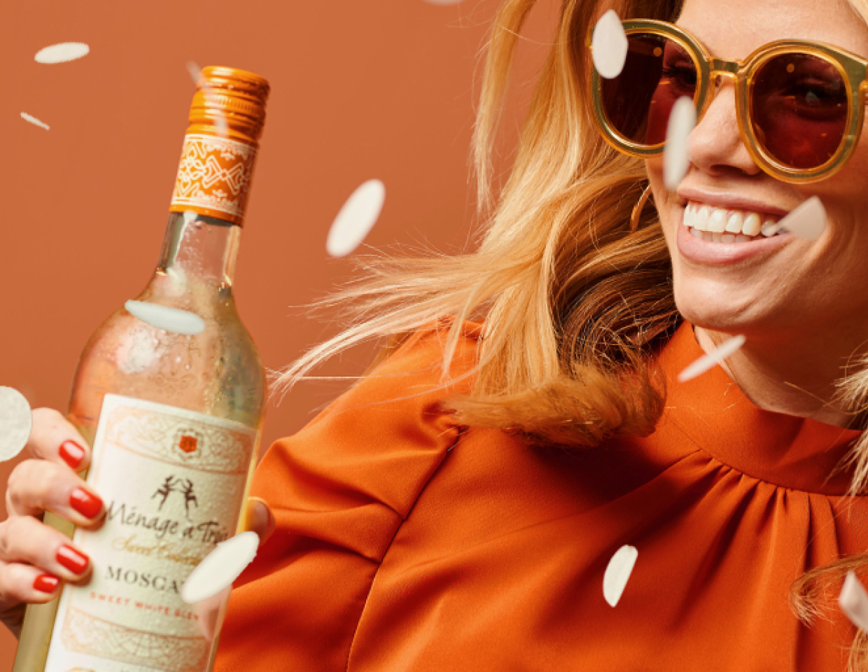 Smiling woman wearing sunglasses and bright orange holding a bottle of Menage a Trois sweet moscato