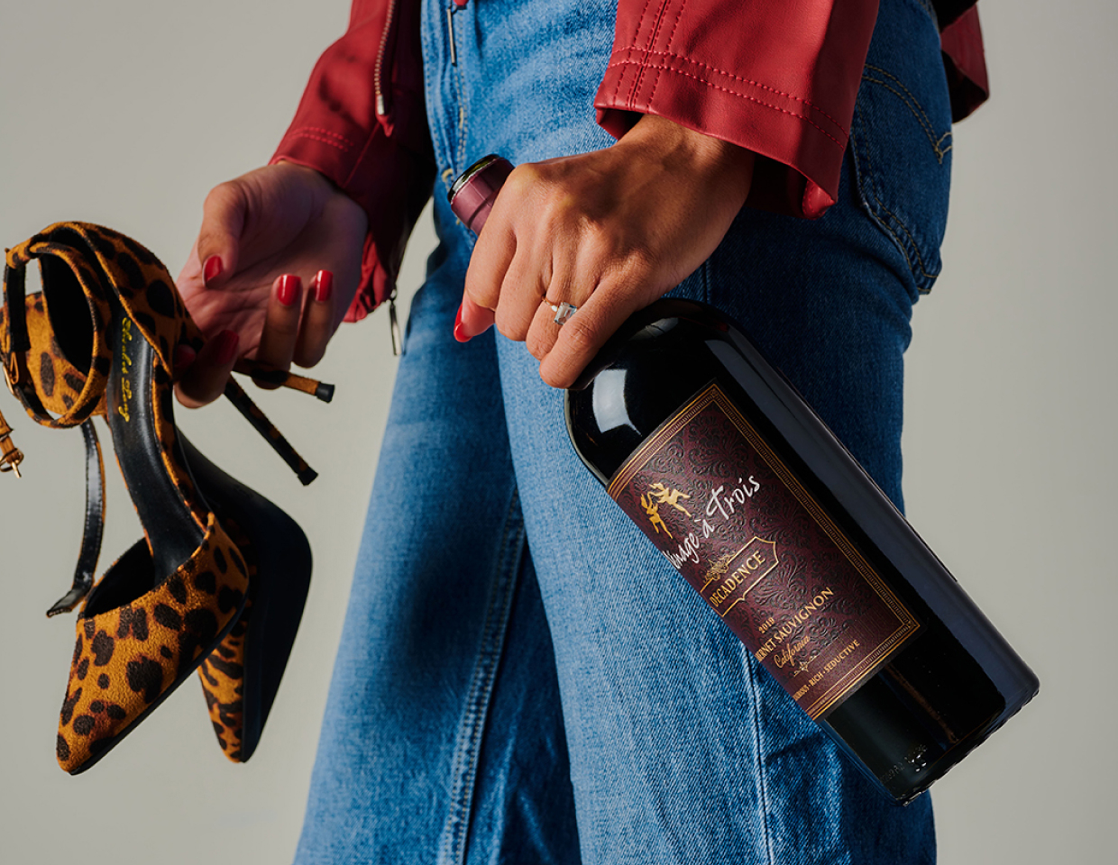 Woman in jeans and red jacket holding leopard print high heels and a bottle of Menage a Trois Decadence.