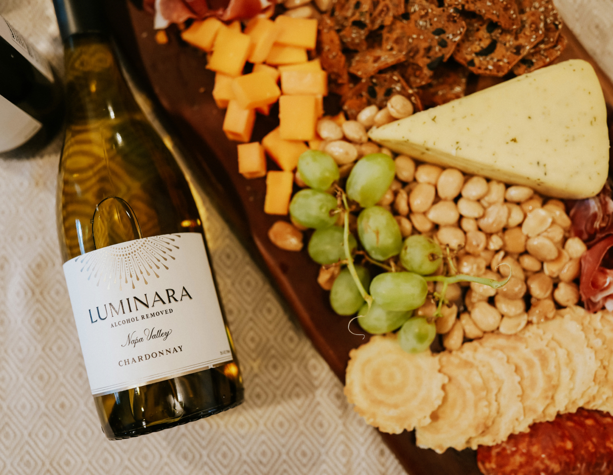 Bottle of Luminara Chardonnay laying down next to a cheese, crackers, nuts, and grapes spread on a wood cutting board.