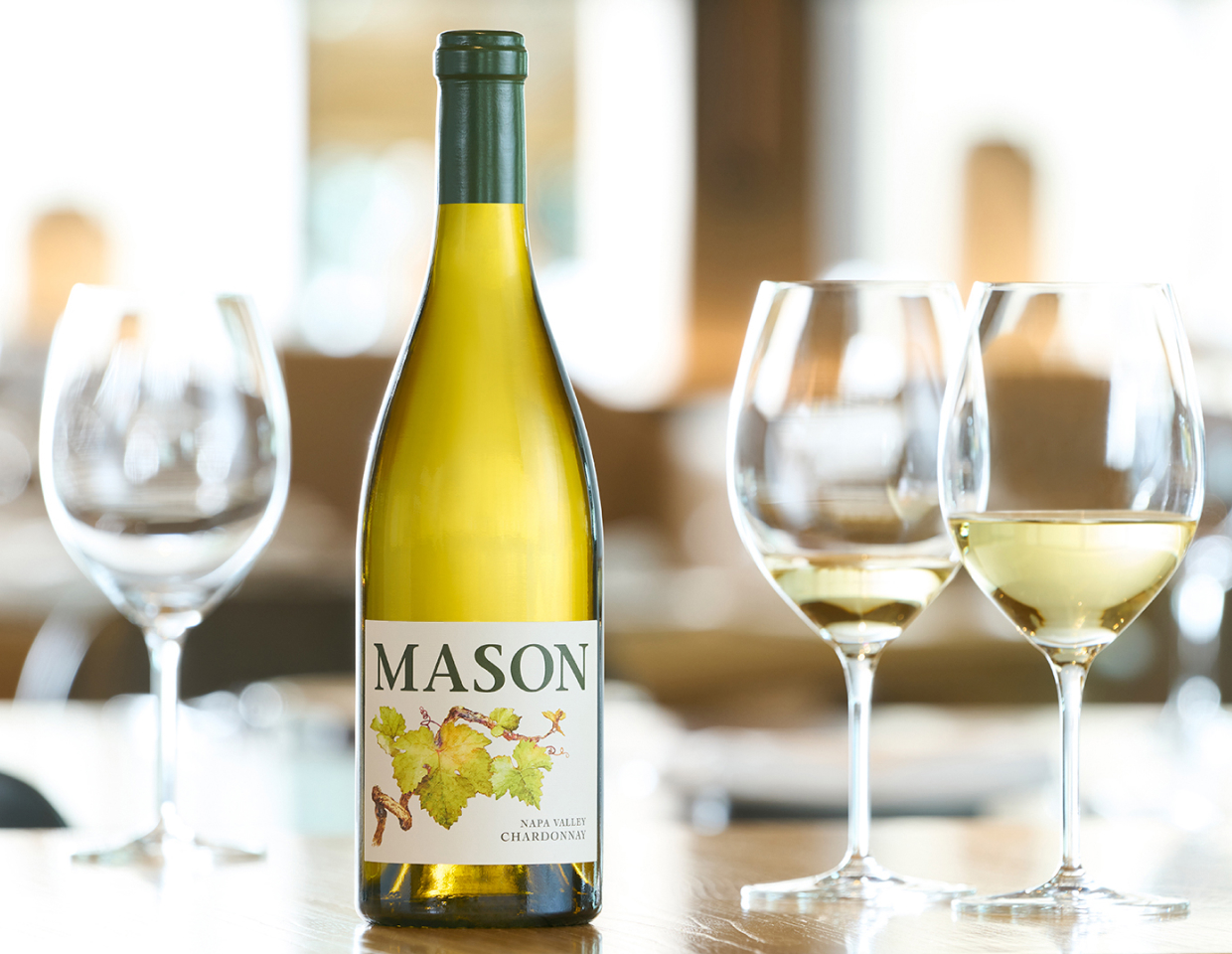 Bottle and glasses of Mason chardonnay and glasses of wine on a wood table in a brightly lit room.
