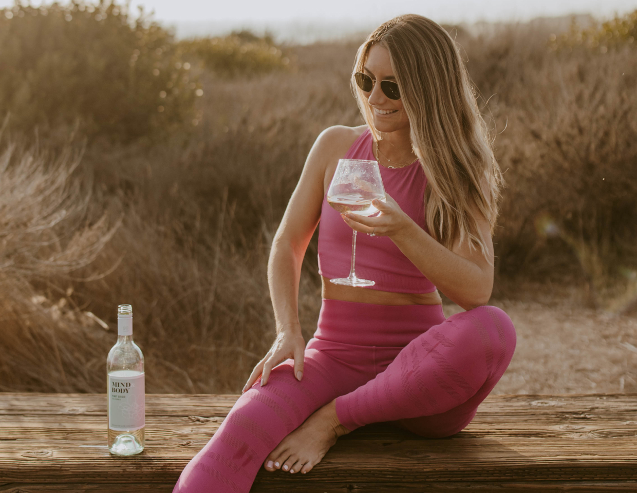 Women sitting on a bench in a beautiful beachy setting wearing a pink athletic outfit holding a glass of Mind & Body Pinot Grigio