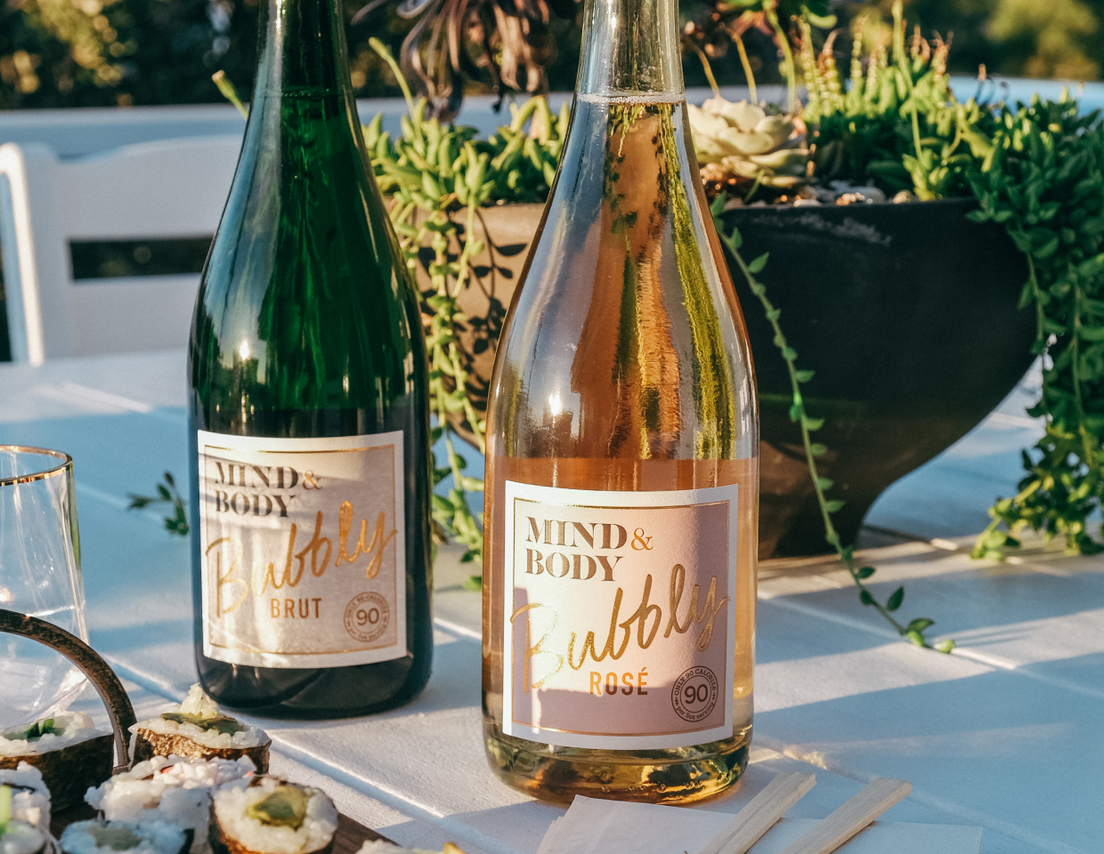 Bottle of Mind & Body bubbly brut and Mind & Body bubbly rose on a table outside next to a plate of sushi and lush green plant.