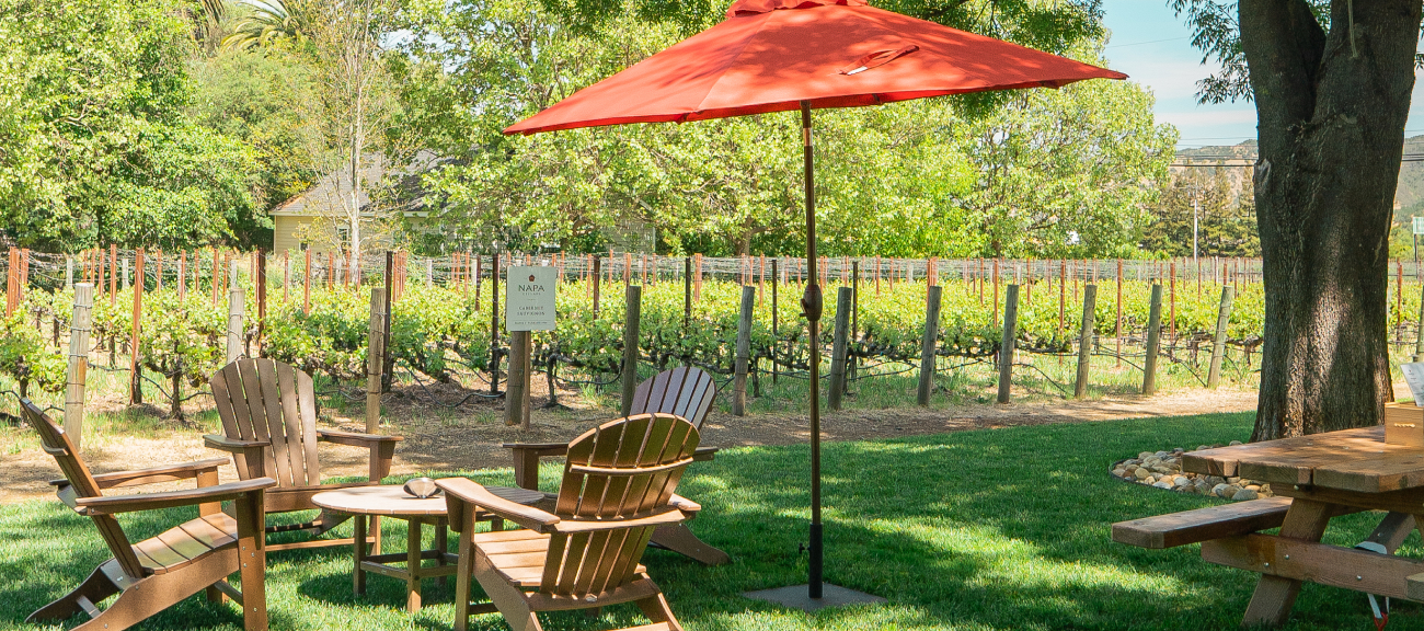 Outdoor seating under a red umbrella in a lush green setting at the Napa Cellars tasting room.