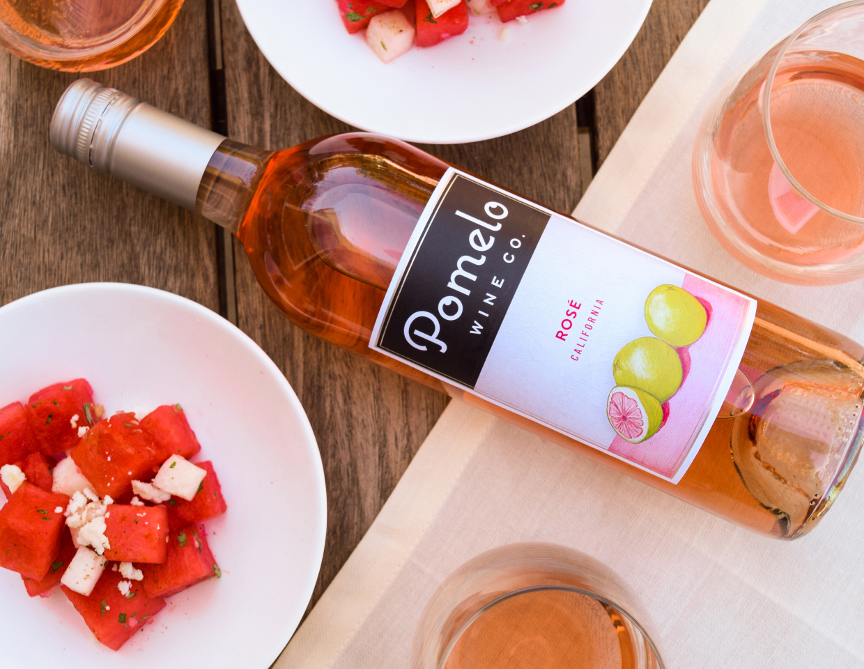 Bottle of Pomelo rose in on a table surrounded by wine glasses and bowls of watermelon and goat cheese salad