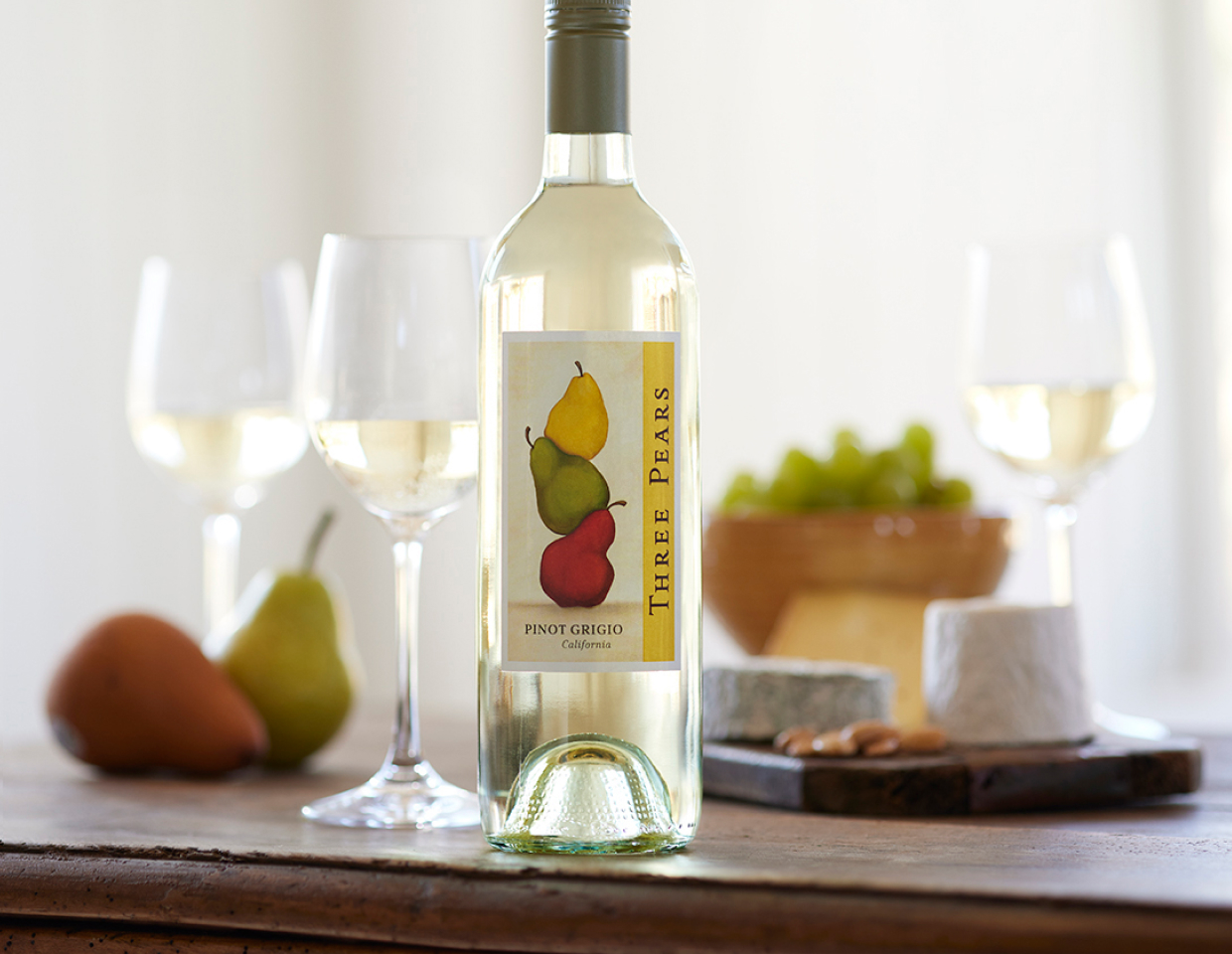 Bottle of Three Pears pinot grigio on a table with glasses of wine, cheese, and fruit