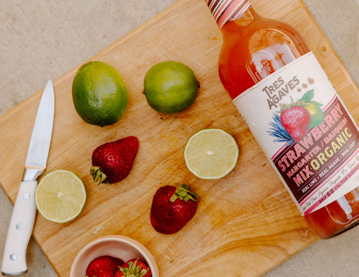 Bottle of Tres Agaves strawberry margarita mix on a cutting board next to limes and strawberries