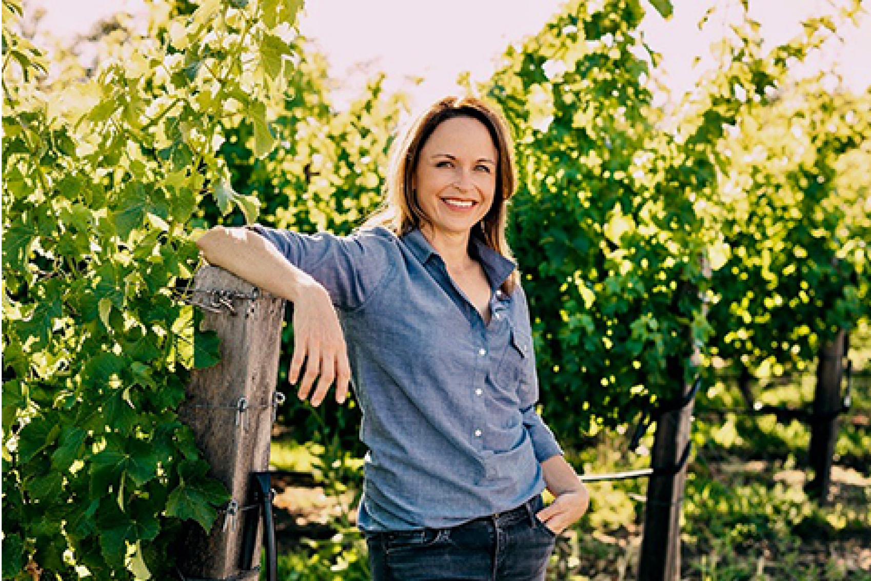 Winemaker of ZIATA wines Jennifer Williams leaning on a fence and smiling by the vines in the sunlight vineyards