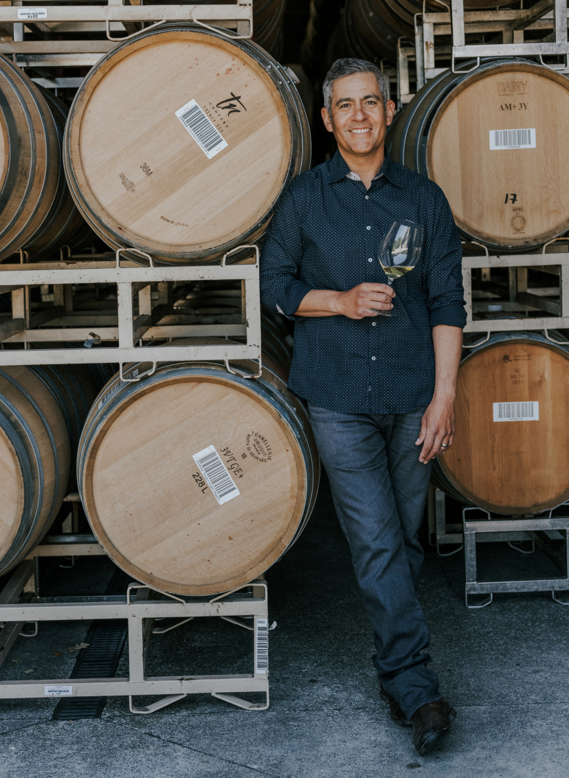 Neyers winemaker Tadeo Borchardt posing in front of wine barrels with a glass of wine