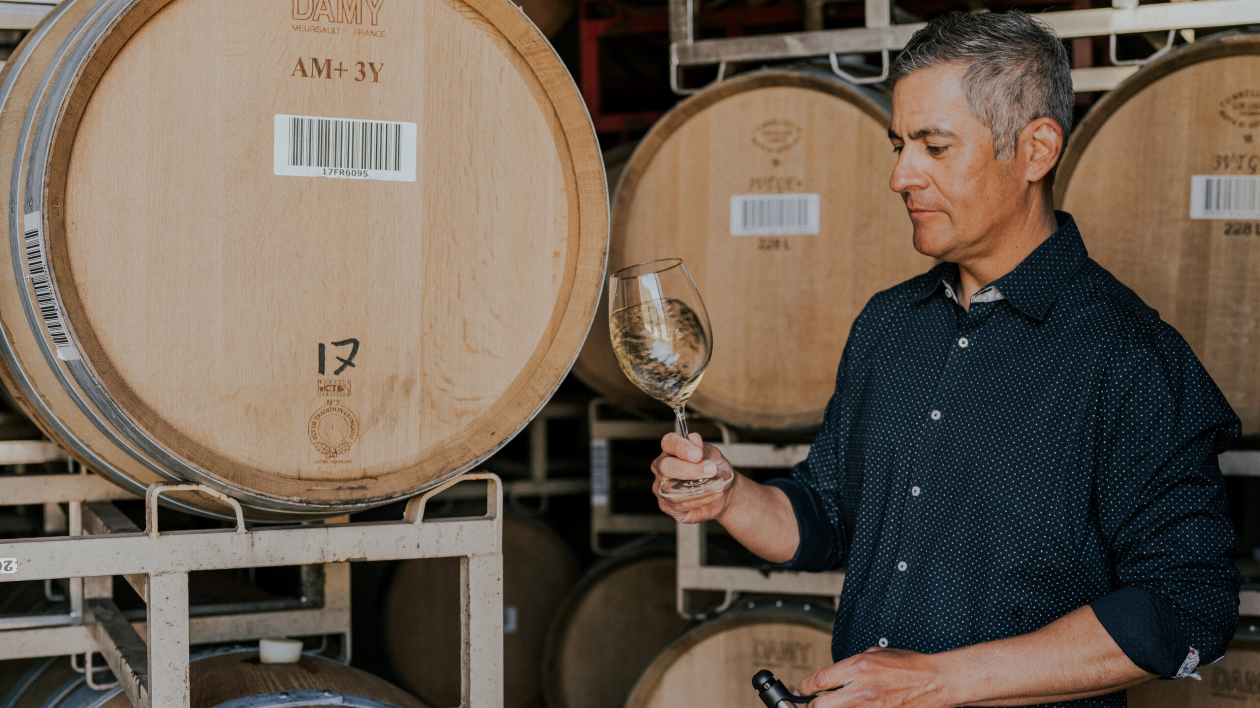 Neyers winemaker Tadeo Borchardt swirling wine in a glass in front of rows of wine barrels
