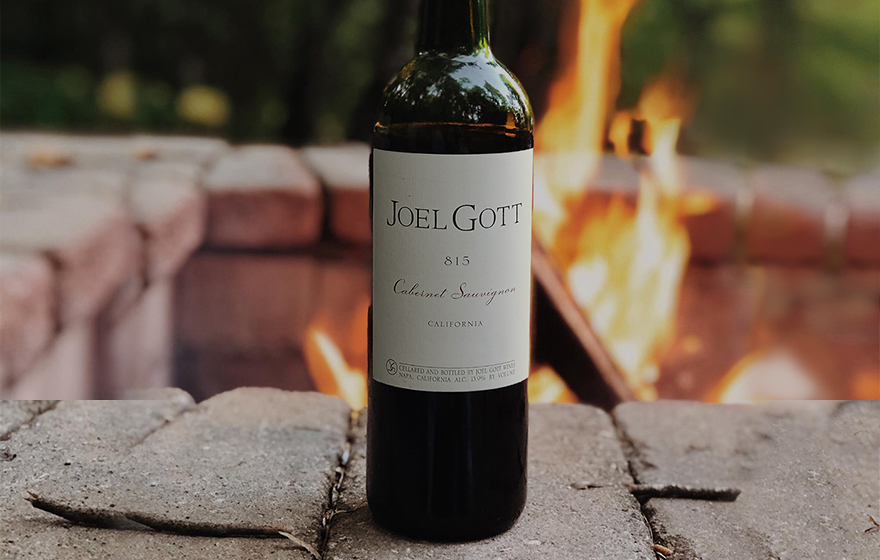 Bottle of Joel Gott cabernet sauvignon on the edge of a fire pit with green trees in the background