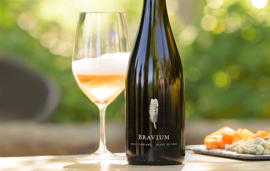 Bottle and glass of Bravium blanc de noir on a table outside with a plate of snacks