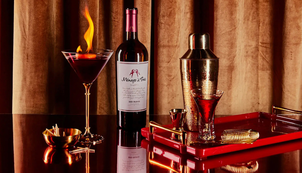 naughty or nice wine cocktail in a martini glass with a bottle of menage a trois red blend next to a shiny cocktail shaker on a reflective surface