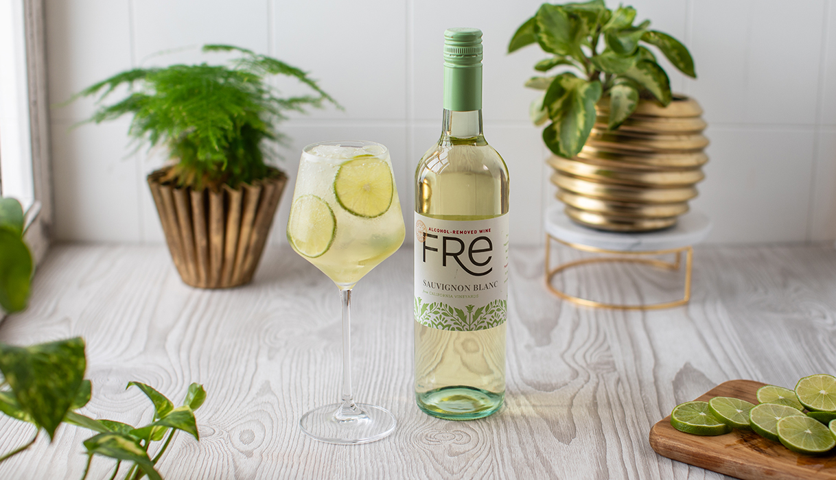 FRE lime spritz mocktail in a wine glass next to a bottle of FRE alcohol-removed sauvignon blanc on a white counter with bright green plants in gold pots