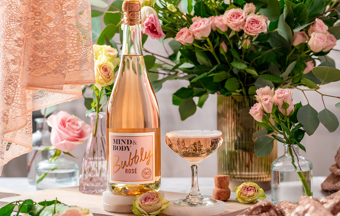 bottle and glass of Mind & Body bubbly rose on a table with beautiful pink roses all around