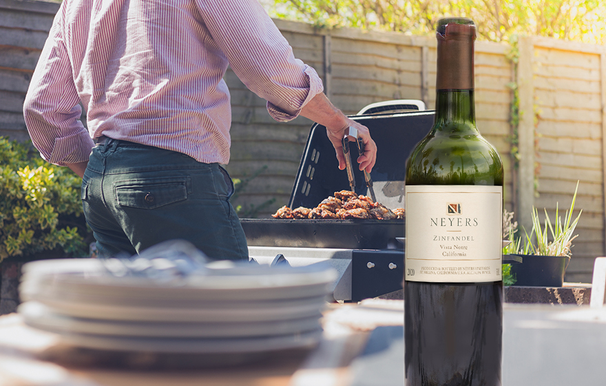 man grilling steak outdoors with a bottle of Neyers zinfandel in the foreground next to a stack of plates