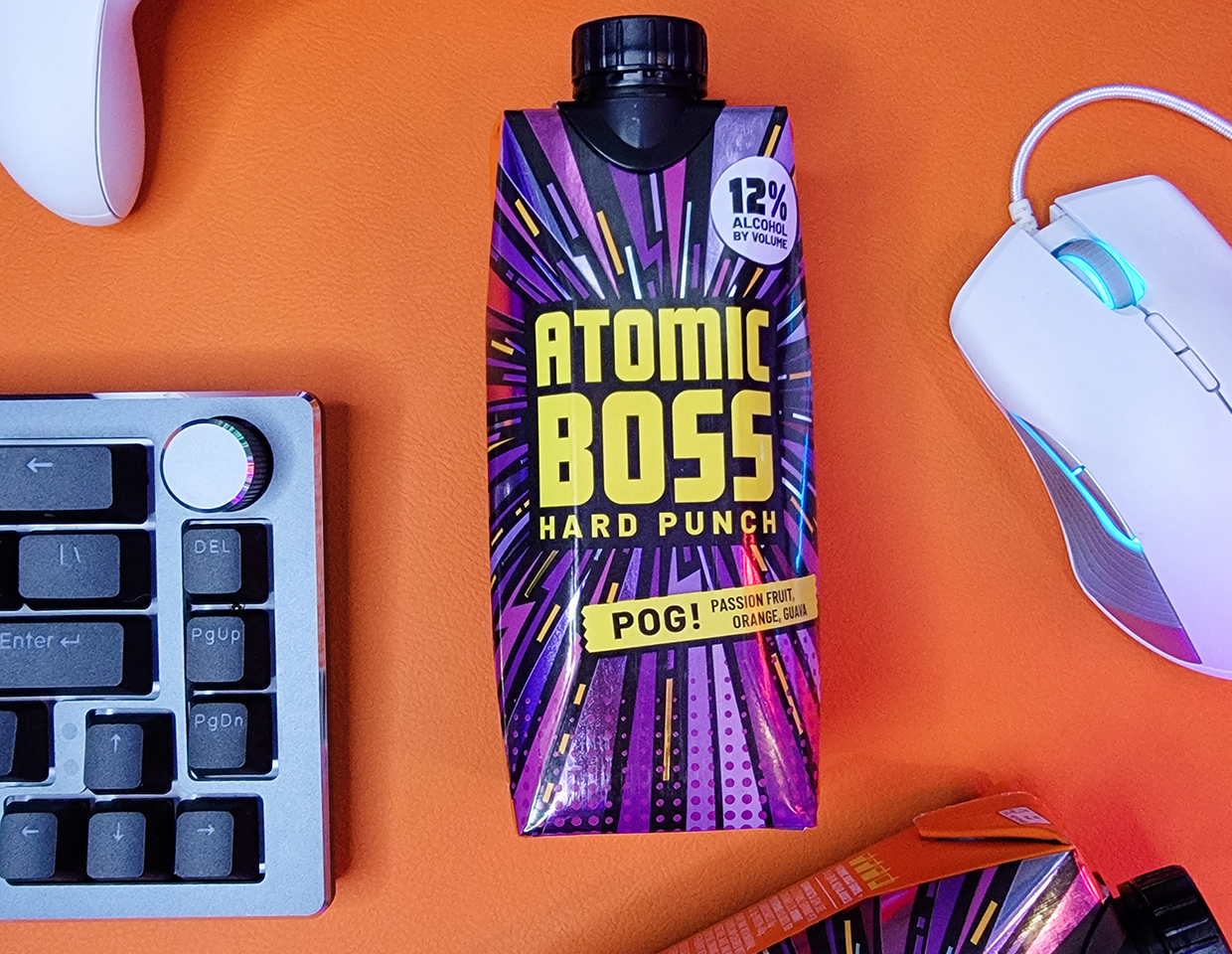 Box of Atomic Boss Hard Punch, POG! flavored: passionfruit, orange, and guava on an orange desk next to a keyboard and mouse.