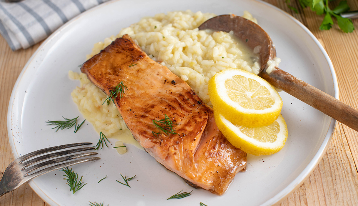A white plate with a seared salmon fillet topped with dill and slices of lemon on a bed of creamy risotto being served with a wooden spoon