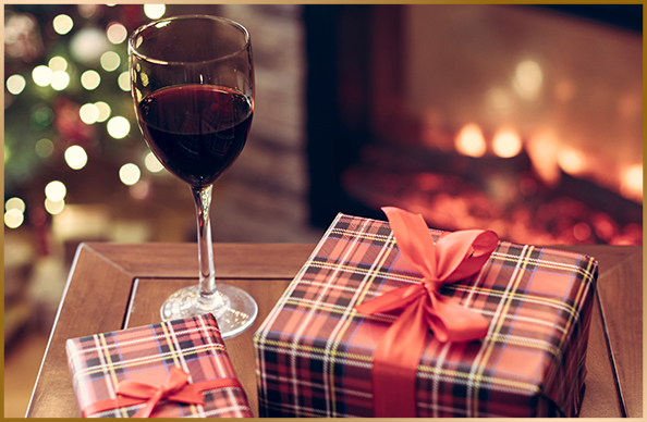 glass of red wine with wrapped holiday gifts in front of a fireplace with a roaring fire and a tree decorated with white lights