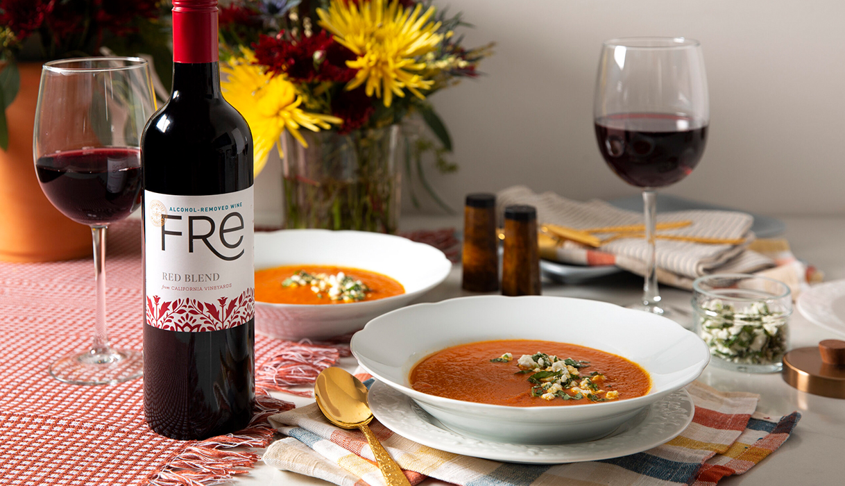 Two bowls of Turmeric roasted tomato soup with goat cheese, next to a bottle and glasses of FRE alcohol-removed red blend with a vase of fall flowers in the background.