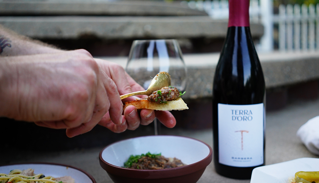 person using a spoon to put bagna cauda on a piece of bread with a bottle of Terra d'Oro Barbera and a glass of wine in the background
