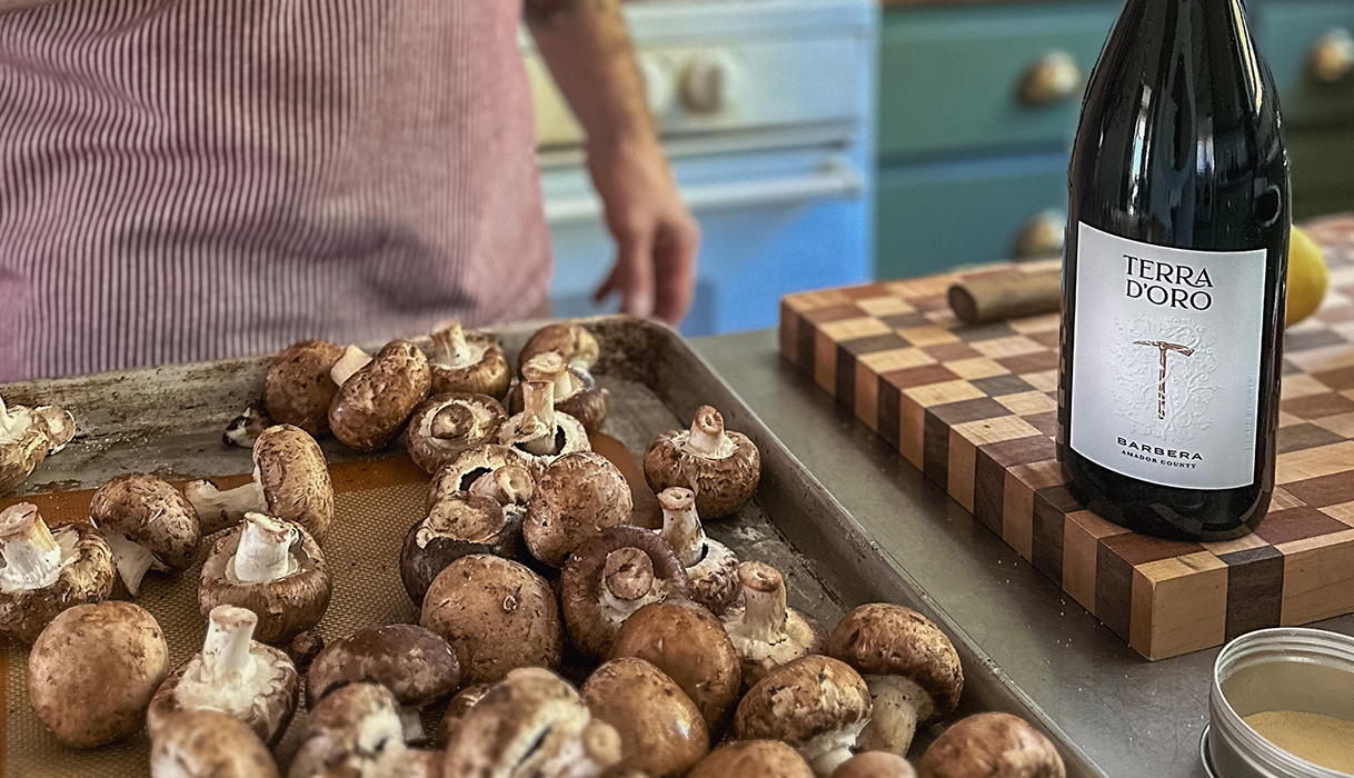 man standing behind a baking tray full of mushrooms next to a bottle of Terra d'Oro Barbera on a butcher block.