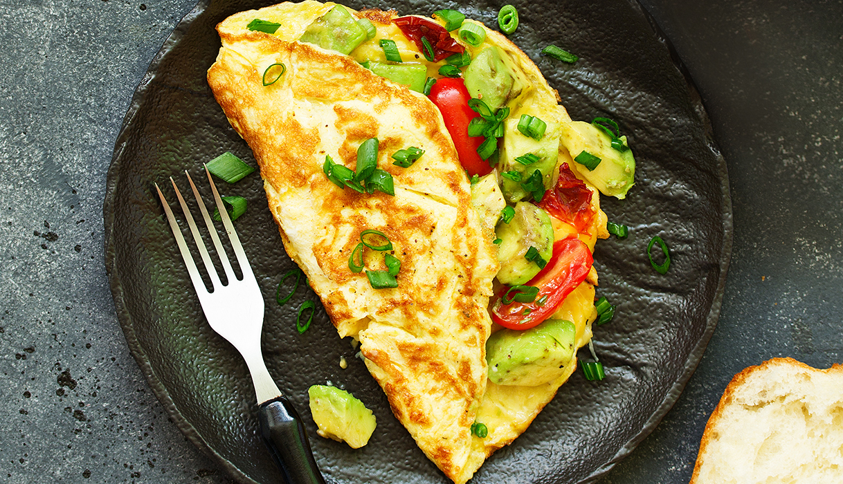 a California omelet bursting with fresh veggies on a black plate with a fork and a piece of bread nearby.