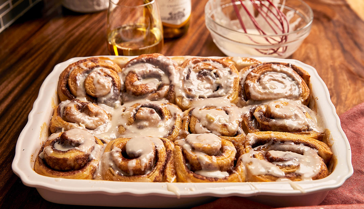 Tray of cinnamon rolls covered with gooey eggnog frosting, with a bottle and glass of Sutter Home chardonnay in the background.