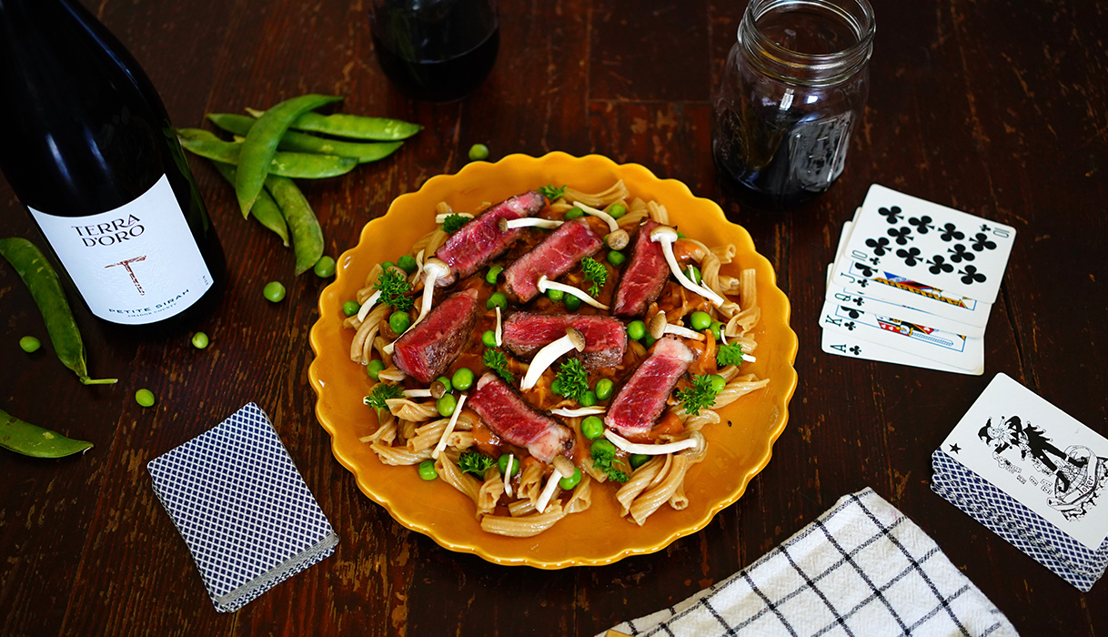 A plate of beef stroganoff next to a bottle of Terra d'Oro Petite Sirah surrounded by peas and playing cards.