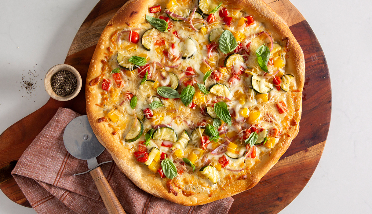 A delicious-looking ranch vegetable pizza on a cutting board with a pizza cutter on a napkin nearby and some fresh ground black pepper.