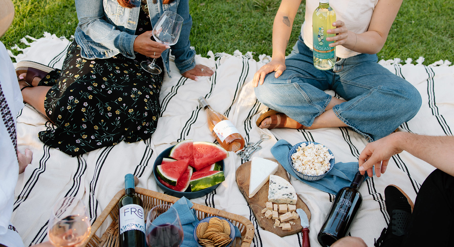 Four people sitting on a picnic blanket on fresh green grass with bottles of various One Stop Wine Shop Wines, a plate of watermelon, a bowl of popcorn, and a plate of cheeses.