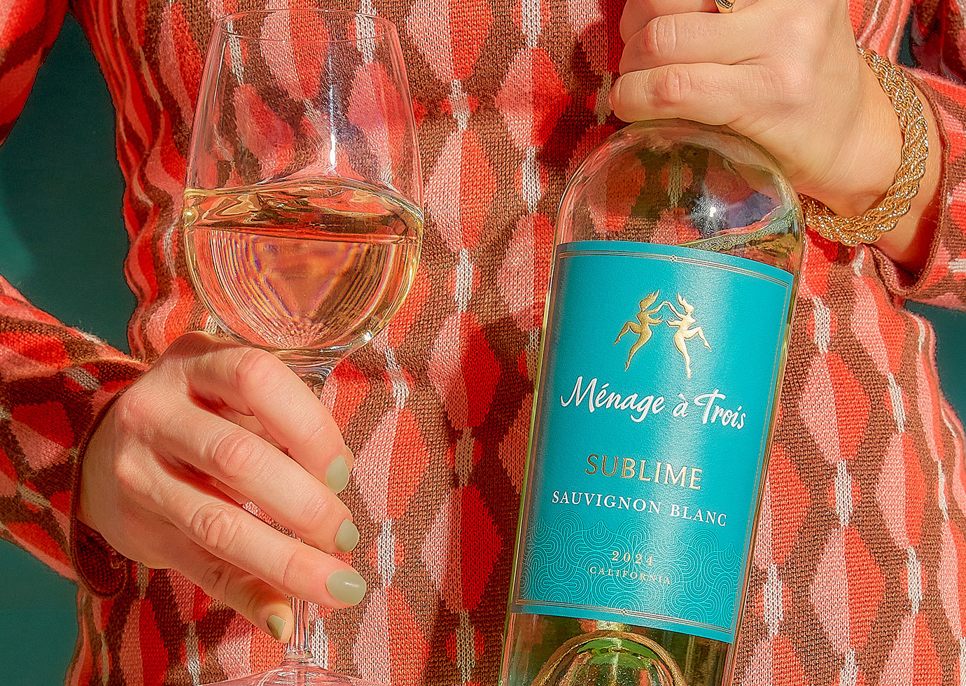 A woman in a bright orange patterned dress holding a wine glass and bottle of Menage a Trois Sublime sauvignon blanc.