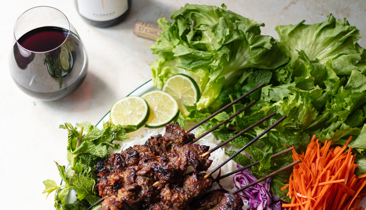 A plate of lemongrass beef skewers on a lush bed of lettuce, mint, limes, and cucumbers, next to a bottle and glass of Terra d'Oro Barbera.