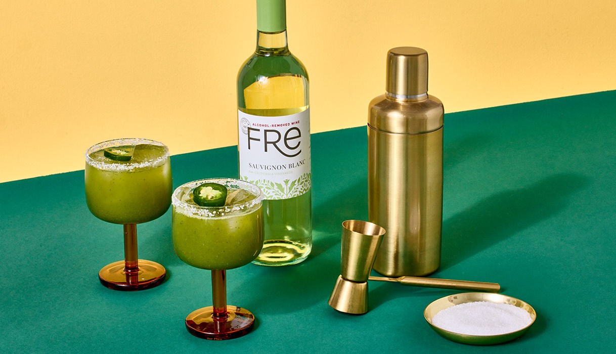 Two glasses of FRE Green dream mocktails, next to a bottle of FRE alcohol-removed sauvignon blanc and a gold cocktail shaker, on a green surface with a yellow background. Try this unique mocktail.