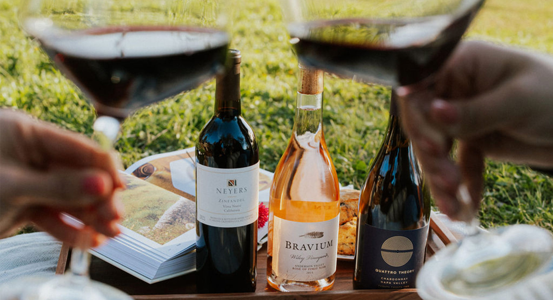 Two people clinking glasses of red wine with a bottle of Neyers zinfandel, Bravium rose of pinot noir, and Quattro Theory chardonnay in the background, sitting on a picnic blanket over green grass. Shop these and more seasonal favorites from One Stop Wine Shop.