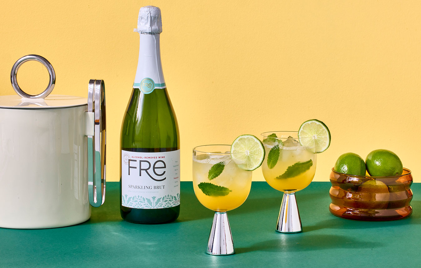 Glasses of FRE mango mojitos garnished with lime circles next to a bowl of limes, and ice bucket, and a bottle of FRE alcohol-removed sparkling brut. Get this summer mocktail recipe and more.