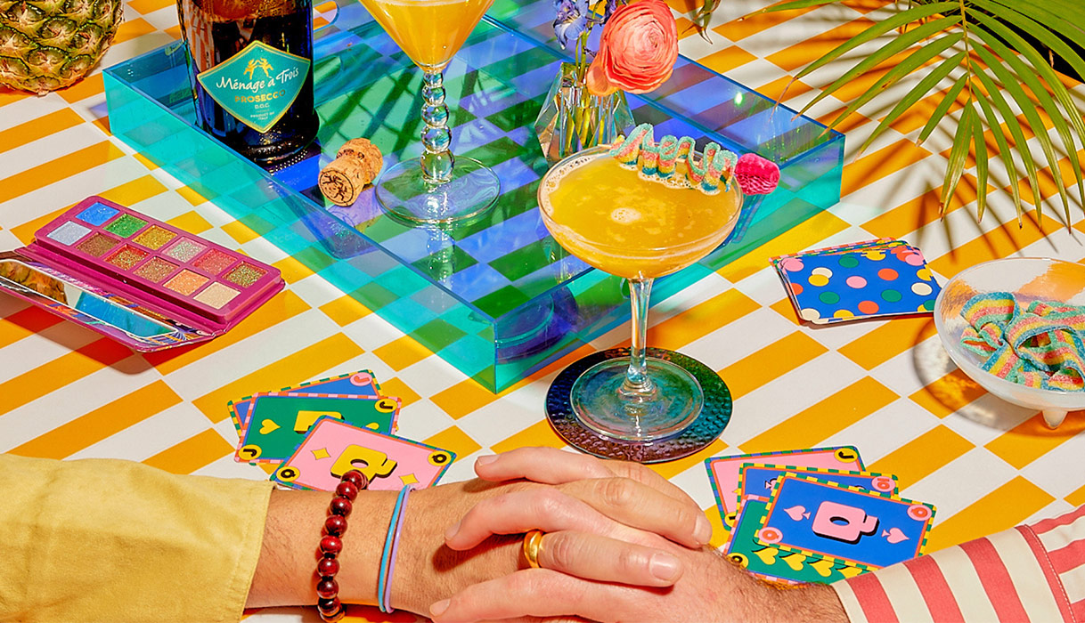 two Kiss & Tell cocktails, made with Menage a Trois prosecco, on a yellow checked tablecloth with two people holding hands.