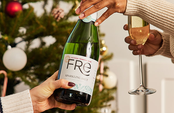 One person handing another a bottle of FRE alcohol-removed sparkling brut in front of a Christmas tree decorated with glass bulbs and candy canes. Buy this and more alcohol-removed wines from One Stop Wine Shop.