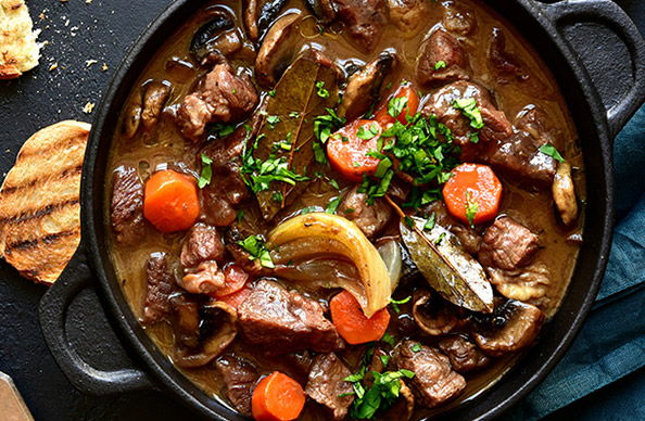 A pot of boeuf bourguignon chock full of meat and vegetables on a countertop next to some toasted pieces of bread. Get this recipe and more from One Stop Wine Shop.