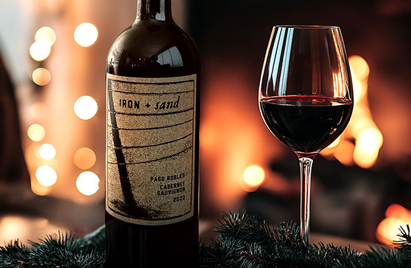 a bottle and glass of Iron + Sand Paso Robles Cabernet Sauvignon by a roaring fire with festive lights in the background and greenery around the bottle. Buy this and more seasonal favorite wines from One Stop Wine Shop.