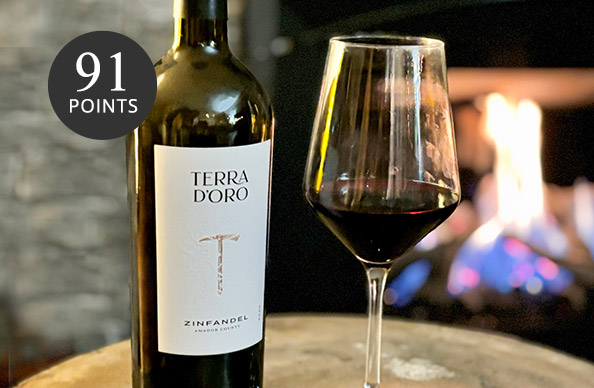 A bottle and glass of Terra d'Oro Zinfandel by a roaring fireplace. This wine is rated 91 points, by this and more top-rated wines from One Stop Wine Shop.