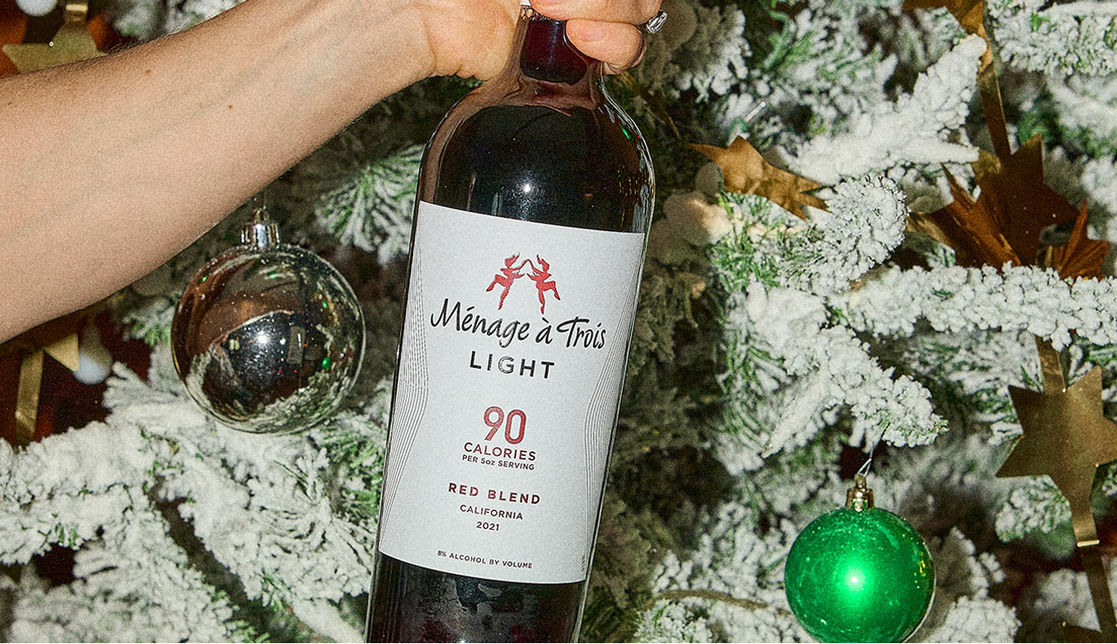 person holding a bottle of Menage a Trois light red blend, 90 calories per 5 oz serving, in front of a decorated Christmas tree. Buy this and more at One Stop Wine Shop.
