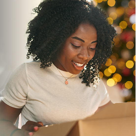 A woman happily unboxing a One Stop Wine Shop wine club shipment with a Christmas tree in the background. Give the gift of wine club.
