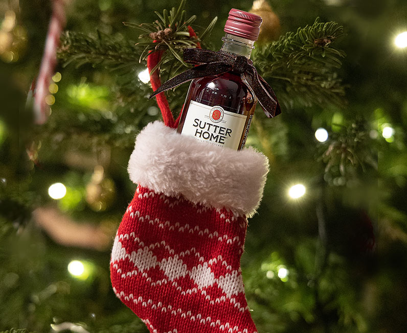 A mini bottle of Sutter Home in a tiny stocking hanging on a tree with lights. Shop mini wine bottles at One Stop Wine Shop.