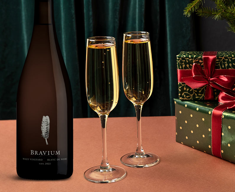 A bottle of Bravium Blanc de Noirs next to two filled champagne glasses and some festive wrapped presents. Buy this and more sparkling wines at One Stop Wine Shop.